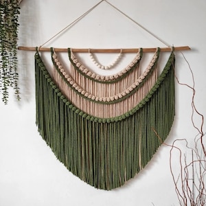 Op de afbeelding: Handgemaakte macramé wandhanger met een halfrond ontwerp. Het stuk is gemaakt met olijfgroen, beige en crèmekleurig katoenen koord. Het is opgehangen aan een natuurlijke houten stok en een jute touw, en is ongeveer 60 cm breed.
