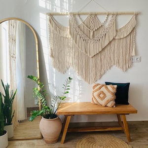 May include: A cream-colored macrame wall hanging with a geometric design is displayed above a wooden bench. A decorative pillow with a diamond pattern and a dark green pillow rest on the bench. A large gold-framed mirror and potted plants are also in the room.