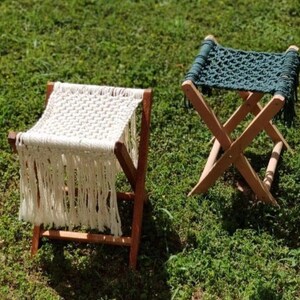 Puede incluir: Dos taburetes plegables de madera con asientos de macramé. Un taburete tiene un asiento de macramé blanco con flecos, y el otro tiene un asiento de macramé verde oscuro.