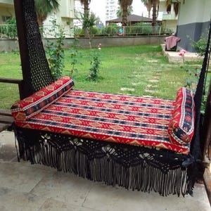 May include: A black hammock swing with a red, black, and gold patterned cushion and two matching pillows. The hammock is hanging from a wooden frame and is surrounded by green grass.