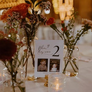 May include: A white card with black calligraphy that reads "When they were 2" with a heart symbol. The card has two photos of children, one boy and one girl, and the text "Karter + Kielle 2-12-22". The card is on a table with flowers in vases.