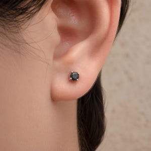 May include: A small, round, dark grey stud earring featuring a single faceted stone held in place by four metal prongs. The earring is positioned centrally on the earlobe, showcasing a simple, circular design with a polished metallic setting.
