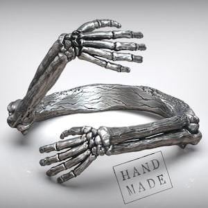 Puede incluir: Un brazalete plateado con forma de brazos esqueléticos abrazándose. El brazalete presenta estructuras óseas detalladas y está marcado con un sello "HAND MADE". El diseño es intrincado y de estilo gótico.