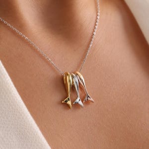 Könnte beinhalten: Eine Halskette mit einer feinen silbernen Kette und drei kleinen, glatten, metallischen Delfinschwanz-Anhängern. Die Anhänger sind in drei verschiedenen Farben gehalten: Gelbgold, Silber und Roségold, alle mit einer glänzenden Oberfläche.