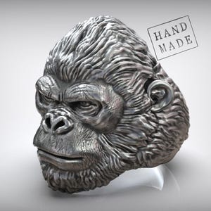 Könnte beinhalten: Ein detaillierter silberner Ring in Form eines Gorillakopfes. Der Ring weist aufwendige Texturen und Gesichtszüge auf, darunter Augen, Nase und Mund. Die Worte "HAND MADE" sind oben rechts gestempelt.
