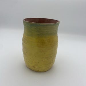May include: A yellow ceramic vase with a green and brown rim. The vase has a slightly rounded shape and a textured surface.