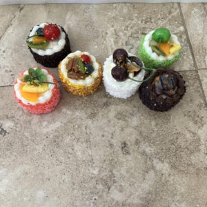 May include: A collection of six miniature decorative cakes. Each cake is round and topped with white frosting and various fruits, nuts, and candies. The cakes have different colored bases, including chocolate, coconut, and sprinkles.