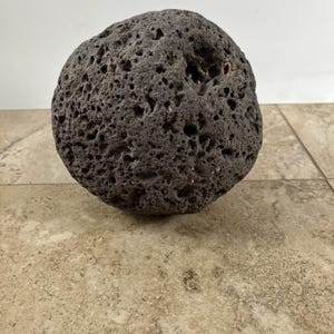 Peut inclure: Une pierre volcanique sphérique de couleur gris foncé avec une texture de surface très poreuse, rugueuse et piquée. La roche présente de nombreux petits trous et des indentations irrégulières sur tout son extérieur, révélant un aspect naturel.