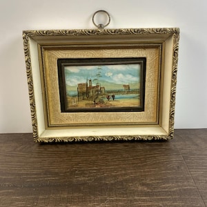 Puede incluir: Pintura de paisaje enmarcada que representa una escena costera con edificios y figuras. La pintura está enmarcada en un marco dorado con un borde blanquecino y un patrón decorativo. Se adjunta un anillo de metal para colgar.