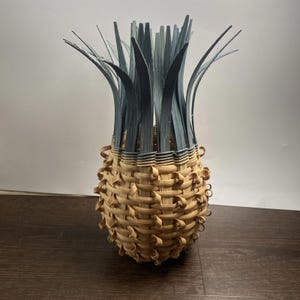 May include: A handcrafted basket resembling a pineapple. The basket features a woven design with natural and blue-gray hues. The top has long, upright blue-gray strands, mimicking the pineapple's crown. The basket's body is a light brown color.