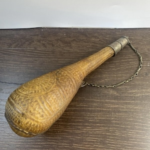 Puede incluir: Una petaca de cuero marrón, alargada, con intrincados patrones en relieve. Tiene una tapa de metal plateado y una cadena. La petaca está sobre una superficie de madera oscura.