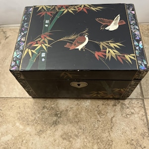 Puede incluir: Caja de joyería rectangular negra con una tapa decorativa con pájaros y bambú pintados. La caja tiene una incrustación de nácar alrededor de los bordes y un ojo de cerradura en forma de corazón.