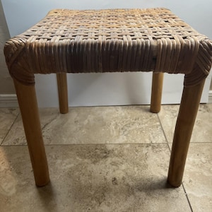 May include: A square stool featuring a woven rattan seat with a natural light brown finish. The stool is supported by four sturdy, cylindrical wooden legs that match the tone of the seat, creating a cohesive and functional piece of furniture.