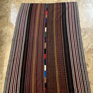 May include: A rectangular textile featuring a vertical striped pattern in shades of brown, black, white, and maroon. The center seam displays a sequence of small, multicolored fabric squares in blue, red, white, green, and black, creating a distinct contrast.