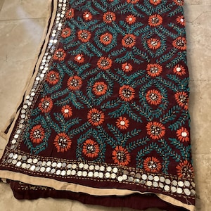 Könnte beinhalten: Ein dunkelmaroonfarbenes Textil mit einem sich wiederholenden Muster aus orangefarbenen gestickten Blumen mit blaugrünen Blattstielen. Der Rand ist mit einer beigen Borte und einer Reihe kreisförmiger silberner Metallspiegel verziert.