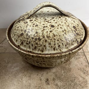 May include: A round ceramic lidded bowl featuring a cream-coloured glaze with dense brown speckles throughout. The lid includes a prominent arched handle, and the entire piece displays a consistent mottled texture across its exterior surface.
