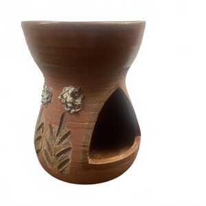 May include: A handmade, brown ceramic oil warmer with a wide top and a rounded base. The warmer features carved leaf designs and two textured floral accents. A large opening is cut into the side.