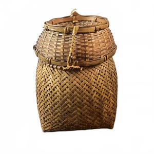 Könnte beinhalten: Ein handgefertigter Korb aus gewebtem Bambus mit Deckel. Der Korb hat einen zylindrischen Boden und eine abgerundete Oberseite, beide aufwendig mit einer natürlichen, hellbraunen Farbe gewebt. Ein gewebter Griff sichert den Deckel. Der Korb wird wahrscheinlich zur Aufbewahrung oder Präsentation verwendet.