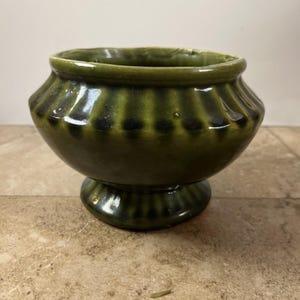 May include: A small, round, olive green ceramic bowl with a glossy finish. The bowl features a wide, fluted rim with vertical indentations around the upper body and a matching fluted pedestal base. The glaze shows darker green shading in the grooves.