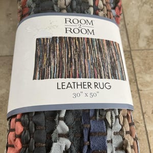 Könnte beinhalten: Ein gerollter Lederteppich mit einer mehrfarbigen Webstruktur aus schwarzen, blauen, grauen und korallenfarbenen Lederstreifen. Das Etikett zeigt ROOM 2 ROOM LEATHER RUG 76,2 cm x 127 cm und eine Grafik des gesamten rechteckigen Teppichmusters.
