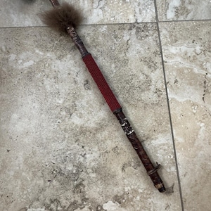 Peut inclure: Un marteau en bois artisanal avec une tête texturée et un long manche décoré. La poignée comporte une section perlée rouge, un accent de fourrure brune et des détails complexes. Le marteau repose sur une surface carrelée.