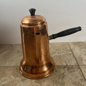 May include: A polished copper coffee pot with a cylindrical body that flares at the base. It features a vertical spout, a removable lid with a small black knob, and a long, horizontal black handle with a ribbed texture for a secure grip.