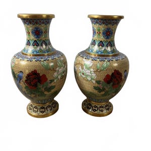 May include: A pair of cloisonné vases featuring a gold-toned background with a fine geometric pattern. The vases are decorated with vibrant red peonies, white magnolia blossoms, and small blue birds. The rims and bases are finished with gold metal.