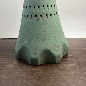 Op de afbeelding: Een groen keramisch decoratief object met een gespikkelde afwerking. Het object heeft een conische vorm met een reeks kleine gaatjes aan de bovenkant en een golvende basis. De kleur is een gedempt groen met roodbruine spikkels.