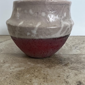 Puede incluir: Un recipiente de cerámica hecho a mano con un esmalte de dos tonos. La parte superior es de color lavanda claro con motas blancas, mientras que la mitad inferior es de color rojo intenso. El recipiente tiene una forma redondeada con un borde ligeramente ensanchado.