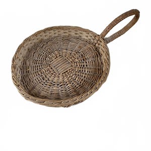 Può includere: Un vassoio rotondo in vimini intrecciato a mano con una finitura naturale color marrone chiaro. Il vassoio presenta una base circolare poco profonda con un motivo intrecciato che si irradia da un dettaglio quadrato centrale e un manico curvo.