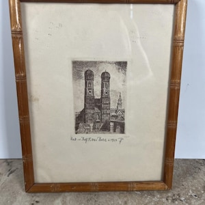 May include: A framed etching featuring the twin-towered Frauenkirche in Munich, set within a light brown bamboo-style wooden frame. The artwork includes handwritten text at the bottom reading Rad. v. Josf F. Osi Probst + 1923.