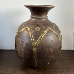 May include: A large, dark brown wooden vase with a wide, rounded body and a narrow neck. The vase is adorned with brass-colored metal bands and rivets, creating a decorative pattern. The vase has a rustic, aged appearance.