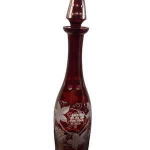 May include: A ruby red glass decanter with a conical stopper. The decanter features etched grape and leaf designs. The stopper has a silver-coloured top with small, evenly spaced dots. The bottle is approximately 30 cm tall.