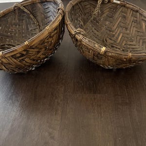 May include: Two woven brown baskets with rope handles. The baskets are round with a wide opening and a tightly woven pattern. The baskets are made of a natural material, likely bamboo or rattan, and have a rustic appearance.