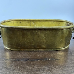 Pode incluir: Uma floreira retangular de latão com cantos arredondados e duas alças curvas. A floreira tem uma aparência desgastada e envelhecida com um tom amarelo dourado. O interior é visível e a floreira está sobre uma superfície de madeira escura.