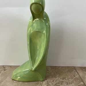 Puede incluir: Una figura de cerámica de color verde claro con una superficie lisa que representa una silueta estilizada con capucha en postura arrodillada o sentada. La escultura tiene un acabado brillante y reflectante con sutiles variaciones de tono.