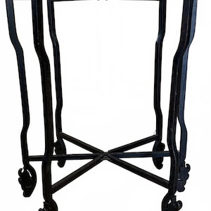 May include: Black metal plant stand with ornate, curved legs and decorative scrollwork. The stand has a square top and a cross-braced base, suitable for displaying plants or decorative items.