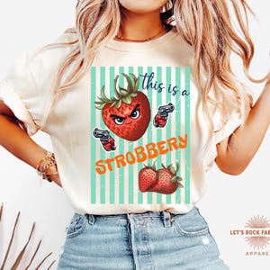 May include: Cream-colored t-shirt featuring a graphic of a strawberry with a stern face holding two guns, with the text "this is a STROBBERY". The design includes a striped background and two strawberries at the bottom.