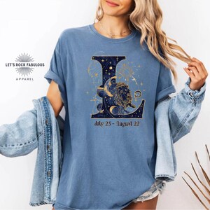 K&ouml;nnte beinhalten: Ein blaues T-Shirt mit einem gro&szlig;en Buchstaben L, der mit einem n&auml;chtlichen Sternenhimmel, einer L&ouml;wenillustration und Mondphasen gef&uuml;llt ist. Unter dem Buchstaben steht der Text July 23 - August 22. Das T-Shirt wird mit einer blauen Jeansjacke getragen.