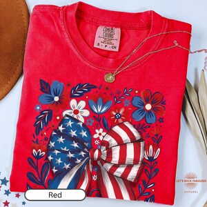 Boho floral American flag bow t-shirt on Comfort Colors 1717 unisex tee. Patriotic 4th of July shirt for women featuring red white and blue bow with floral design. Available in sizes S, M, L, XL, 2XL, 3XL, 4XL and multiple color options including ivory, white, navy, pepper, blossom, and chambray. Perfect America 250th Anniversary tee, Independence Day outfit, summer BBQ, and casual Americana style.