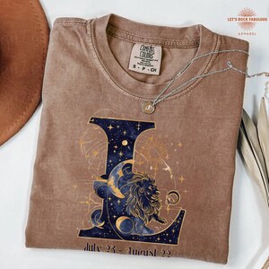 K&ouml;nnte beinhalten: Ein braunes Kurzarm-T-Shirt mit einem gro&szlig;en Buchstaben L, der mit einem dunkelblauen Sternenhimmel, einer Mondsichel und einer L&ouml;wenillustration gef&uuml;llt ist. Unter dem Buchstaben steht der Text July 23 - August 22. Eine silberne Halskette liegt auf dem Shirt.