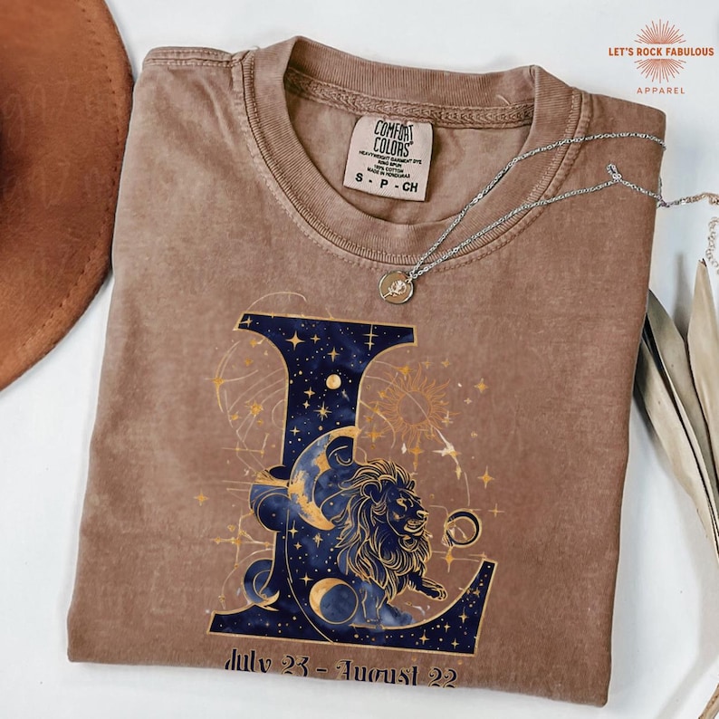 K&ouml;nnte beinhalten: Ein braunes Kurzarm-T-Shirt mit einem gro&szlig;en Buchstaben L, der mit einem dunkelblauen Sternenhimmel, einer Mondsichel und einer L&ouml;wenillustration gef&uuml;llt ist. Unter dem Buchstaben steht der Text July 23 - August 22. Eine silberne Halskette liegt auf dem Shirt.