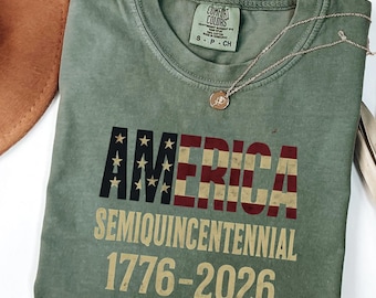 May include: A moss green t-shirt with the word "AMERICA" in a patriotic design, with the American flag incorporated into the letters. Below, it reads "SEMIQUINCENTENNIAL 1776-2026". A gold necklace with a pendant is draped across the shirt.