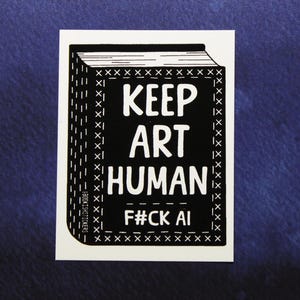May include: A rectangular sticker featuring a black and white illustration of a book. The cover displays the text KEEP ART HUMAN and F#CK AI in white capital letters, surrounded by a decorative border of small white crosses on a black background.