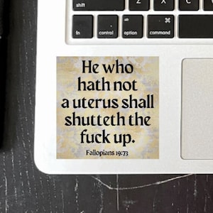 May include: A square sticker with the text "He who hath not a uterus shall shutteth the fuck up." in black font on a textured background. The sticker is on a laptop, with a black pen to the left.
