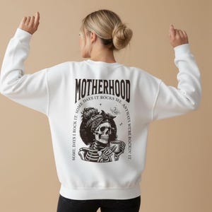 Pu&ograve; includere: Felpa bianca con una grafica sul retro raffigurante uno scheletro con un foulard che tiene una tazza. La parola "MOTHERHOOD" &egrave; sopra la grafica, con il testo "SOME DAYS I ROCK IT. SOME DAYS IT ROCKS ME. ANYWAYS WE'RE ROCKIN' IT."