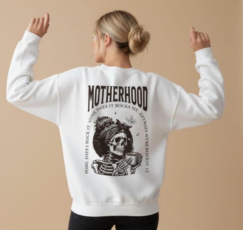 Pu&ograve; includere: Felpa bianca con una grafica sul retro raffigurante uno scheletro con un foulard che tiene una tazza. La parola "MOTHERHOOD" &egrave; sopra la grafica, con il testo "SOME DAYS I ROCK IT. SOME DAYS IT ROCKS ME. ANYWAYS WE'RE ROCKIN' IT."