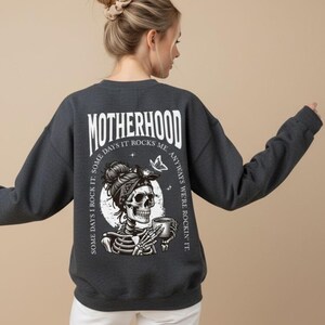 Pu&ograve; includere: Felpa grigio scuro con la scritta "MOTHERHOOD" in lettere bianche. Il retro della felpa presenta una grafica di uno scheletro con una bandana e una tazza, con la scritta "SOME DAYS I ROCK IT. SOME DAYS IT ROCKS ME. ANYWAYS WE'RE ROCKIN' IT."