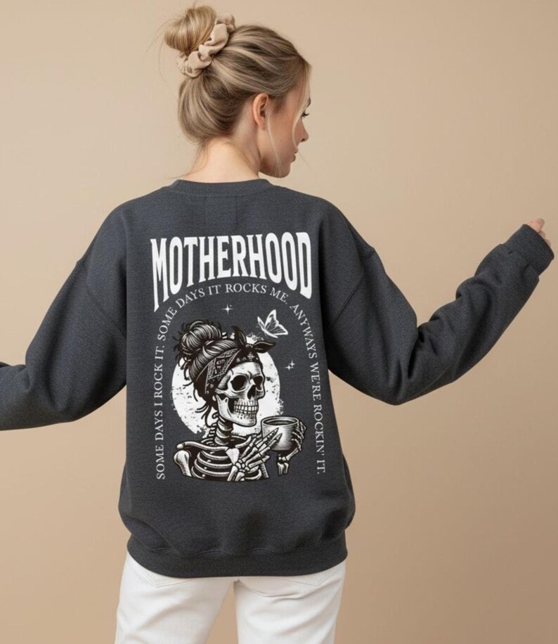 Pu&ograve; includere: Felpa grigio scuro con la scritta "MOTHERHOOD" in lettere bianche. Il retro della felpa presenta una grafica di uno scheletro con una bandana e una tazza, con la scritta "SOME DAYS I ROCK IT. SOME DAYS IT ROCKS ME. ANYWAYS WE'RE ROCKIN' IT."