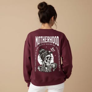 Pu&ograve; includere: Felpa bordeaux con la scritta "MOTHERHOOD" a lettere bianche. Sul retro &egrave; stampata una grafica a scheletro con bandana e tazza di caff&egrave;. Anche il testo "SOME DAYS I ROCK IT. ANYWAYS WE'RE ROCKIN' IT" &egrave; stampato sul retro.